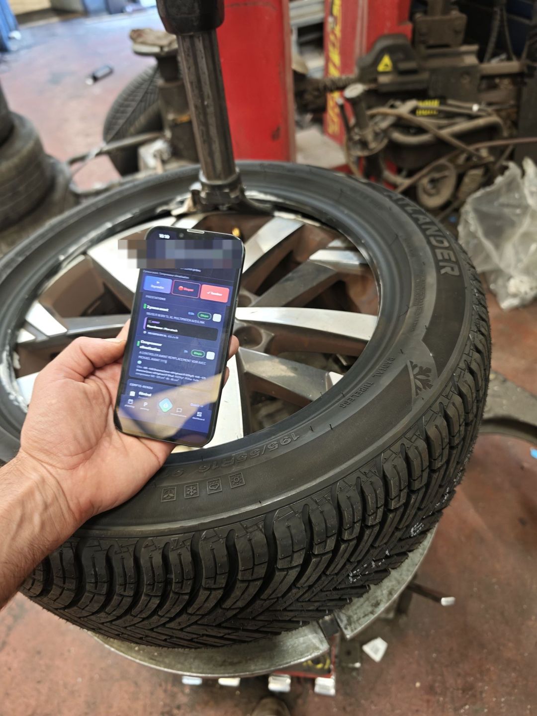 Téléphone avec l'application Axium tenu devant une machine à pneus, pneu hiver monté sur la jante